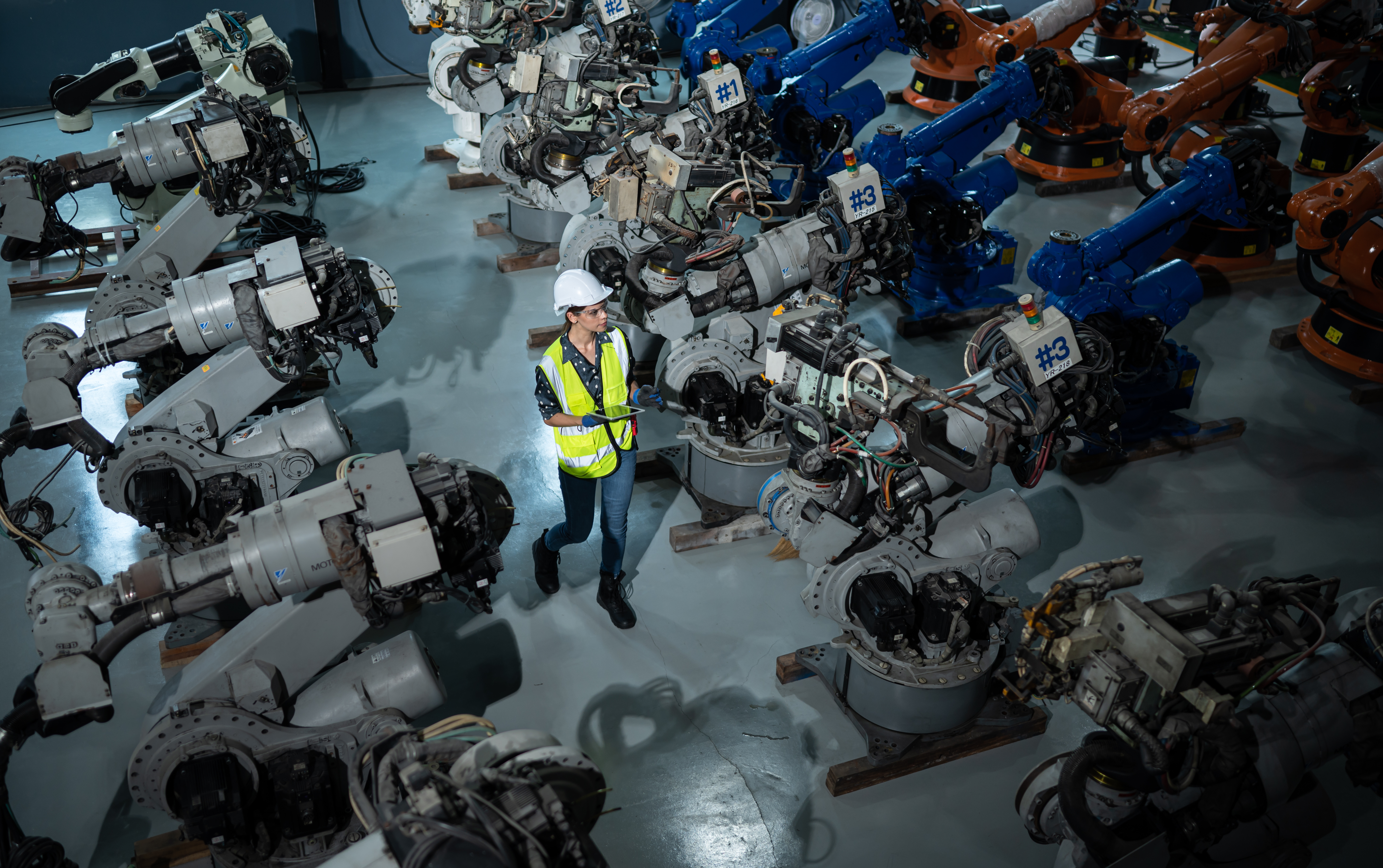 Worker in hard hat and high-visibility vest walking through an automotive robotics line, surrounded by industrial robot arms numbered for tracking.
