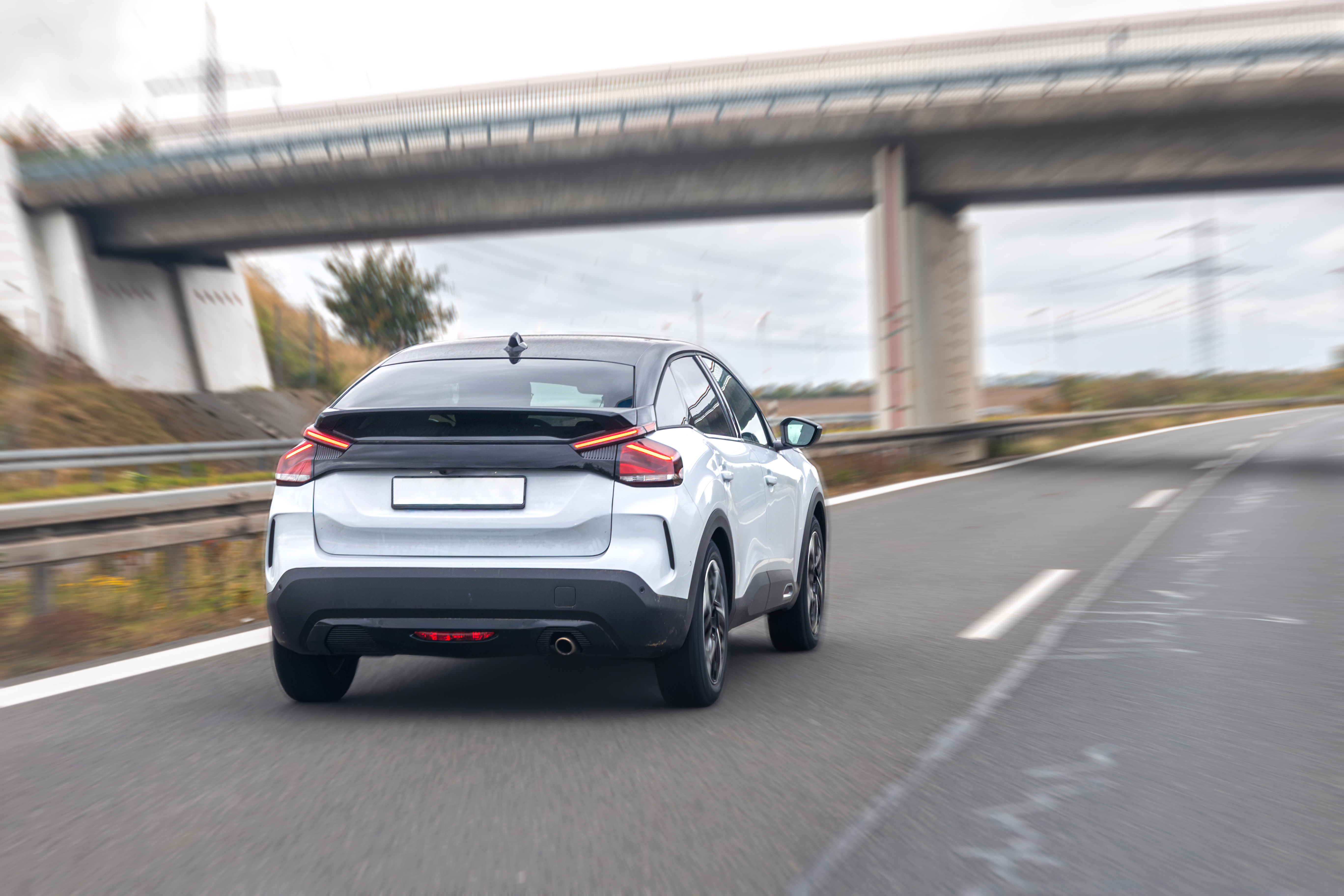 Light-coloured passenger car in motion on a highway, viewed from behind as it passes under an overpass.